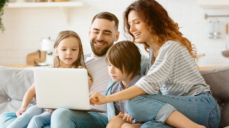 Casal com duas filhas, sentados no sofá e felizes, utilizando notebook